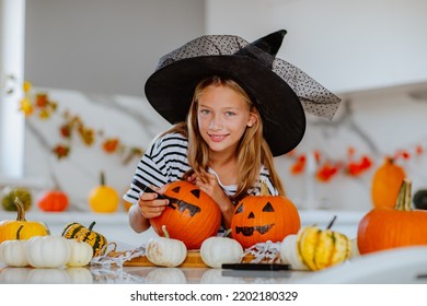 Young pretty girl in black witch hat drawing faces pumpkins for halloween white kitchen  Holiday concept 