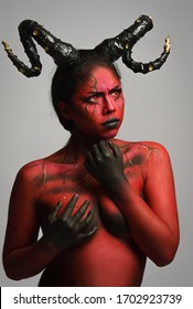 young pretty girl doing body painting characterized by demon with big black horns and red and black body doing poses in photo studio