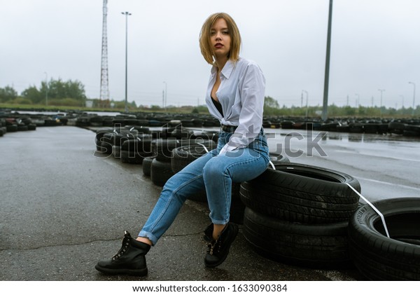 young pretty girl dressed in jeans and white shirt\
standing during autumn little rain outdoors on wet race track, near\
old car tires