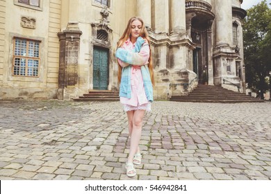 Young Pretty Girl With Extra Long Blonde Hair In Light Pink Dress And Knitted Sweater On The City Streets. Beautiful Fashion Woman Outdoors.