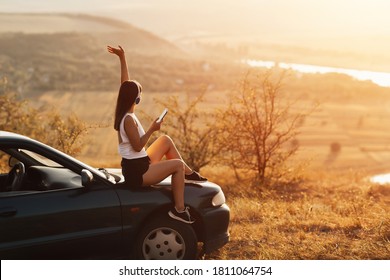 Young Pretty Girl With Hand Up, In Headphones Listening To Music And Singing Sitting On The Hood Of A Car At Sunset. Concept Of Travel, Technology And Happy Life.