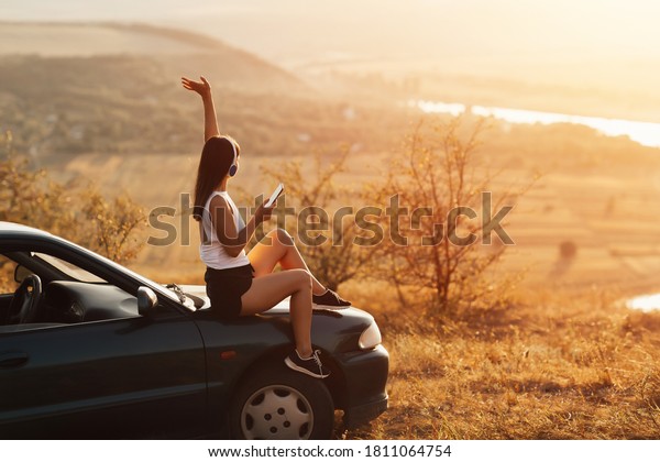 Young pretty girl with hand\
up, in headphones listening to music and singing sitting on the\
hood of a car at sunset. Concept of travel, technology and happy\
life.