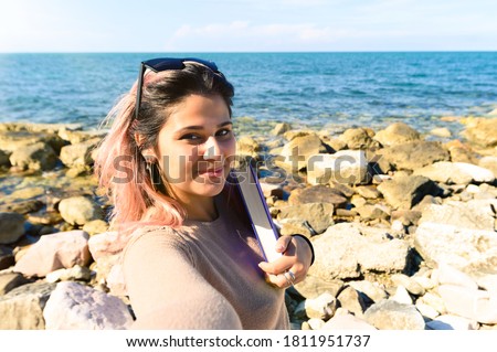 A young pretty girl holds book in her hands on the beach by the sea - Girl in summer hat taking a selfie at the beach - Wild culture lifestyle concept