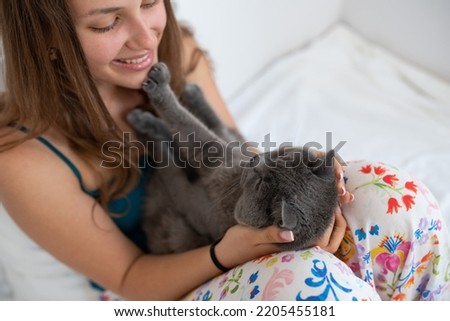 Young pretty girl is playing with her British shorthair cat in her bedroom, his paws touching her face