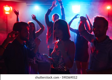 Young pretty girl in shining top dancing with male friends at student disco party and enjoying music with her eyes closed - Shutterstock ID 590369012
