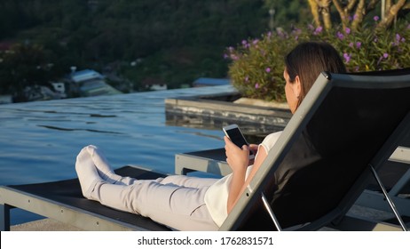 Young Pretty Girl Surfs Internet On Smartphone Resting In Folding Chair At Hotel Side View Close-up