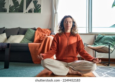 Young pretty healthy mindful woman wearing headphones listening calming music audio podcast sitting on floor at home doing yoga meditation relaxing exercise for mental balance in apartment interior. - Powered by Shutterstock