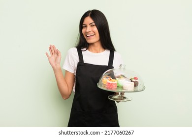 Young Pretty Hispanic Woman Smiling Happily, Waving Hand, Welcoming And Greeting You. Chef With Cakes Concept