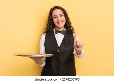 young pretty hispanic woman smiling and looking friendly, showing number one. waiter and tray concept