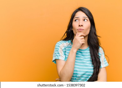 Young Pretty Hispanic Woman Thinking, Feeling Doubtful And Confused, With Different Options, Wondering Which Decision To Make Against Brown Wall