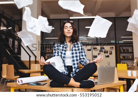 Young pretty joyful brunette woman meditating on table surround work stuff and flying papers. Cheerful mood, taking a break, working, studying, relaxation, true emotions