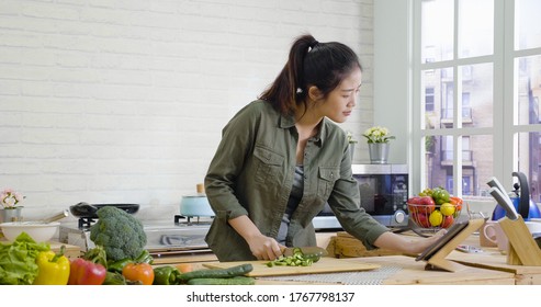 Young Pretty Lady Standing In Modern Kitchen At Home And Cooking. Beautiful Asian Korean Girl Looking At Tablet Computer On Wooden Table Search For Recipe Step While Chopping Cucumber And Vegetable