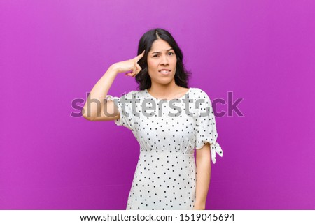 young pretty latin woman feeling confused and puzzled, showing you are insane, crazy or out of your mind against purple wall