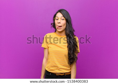 young pretty latin woman looking goofy and funny with a silly cross-eyed expression, joking and fooling around against purple wall