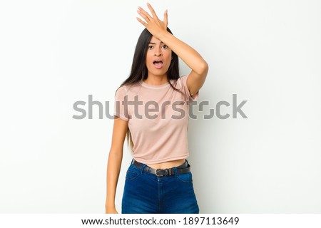 young pretty latin woman raising palm to forehead thinking oops, after making a stupid mistake or remembering, feeling dumb against flat wall