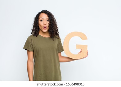young pretty latin woman surprised, shocked, amazed, holding the letter G of the alphabet to form a word or a sentence.