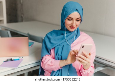 Young Pretty Modern Muslim Woman In Hijab Working On Laptop In Office Room, Education Online, Remote Work Freelancer, Online Conference Chat Conversation Using Smartphone And Earphones