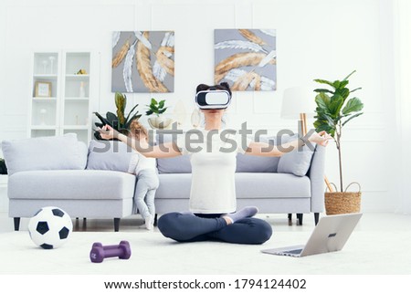 Young pretty mom meditating in lotus yoga position using virtual reality goggles while her daughter watches cartoons at home on background.