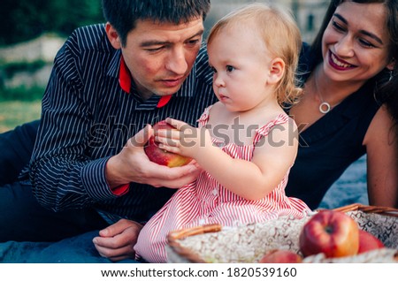 Young pretty mother, father and their little cute daughter with red apple at blanket on picnic
