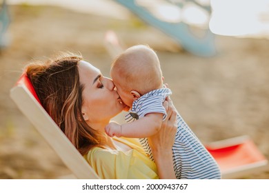 Junge hübsche Mutter in gelbem Sommerkleid mit rotem Strandstuhl küssen ihren Neugeborenen Jungen. 