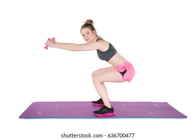 Young, Pretty Natural Looking Girl In Sport Bra And Pink Shorts Showing How To Do Proper Squat Exercise With Dumbbells On The Mat. Full Body Studio Shot, Pure White Background. 
