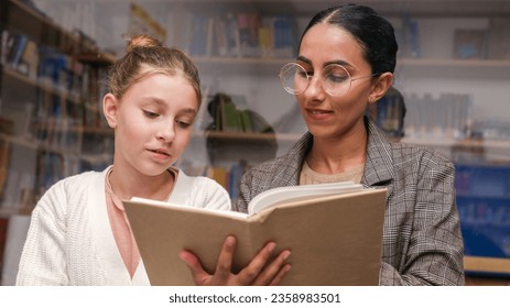 Young pretty Oriental teacher in glasses helps with reading to a student of a blond girl, holding a book, reading in the school library. Back to School. Learning and Education Assistance and concrpt - Powered by Shutterstock