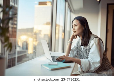 Young pretty pensive Asian professional business woman employee or student working on laptop in corporate office sitting at desk looking at window thinking of new ideas or dreaming, contemplating. - Powered by Shutterstock