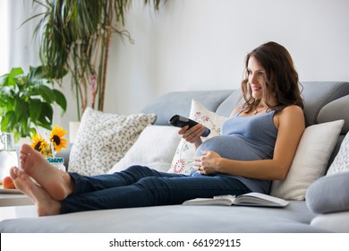 Young Pretty Pregnant Woman Watching TV At Home On The Couch, Eating Healthy, Fruits, Lemonade And Flowers On The Table