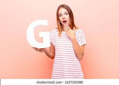 young pretty red head woman surprised, shocked, amazed, holding the letter G of the alphabet to form a word or a sentence.