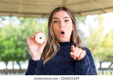Young pretty Romanian woman holding a donut at outdoors surprised and pointing front
