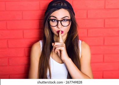 Young Pretty Seductive Hipster Woman Having Fun And Showing Shhh Science, Wearing Black Swag Cap And Clear Glasses, Long Hairs And Bright Make Up. Red Urban Wall Background.