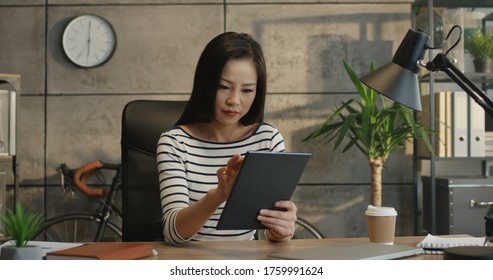 Young Pretty And Smiled Asian Woman, Office Worker, Tapping And Scrolling On The Tablet Device In Her Hands At The Working Table.