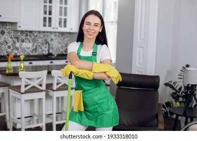 Young pretty smiling brunette Caucasian woman, worker of cleaning company in green uniform, posing on camera with mop and cloth during cleaning up of modern luxury house.