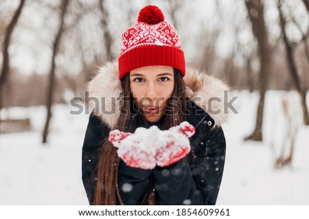 young pretty smiling happy woman in red mittens and knitted hat wearing winter coat, walking in park, playing with snow in warm clothes