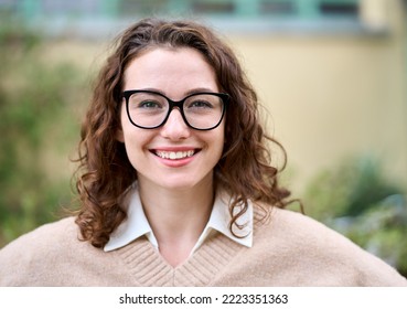 Young pretty smiling professional business woman, happy businesswoman, positive beautiful curly lady wearing glasses standing outdoor on street, looking at camera, front face headshot portrait. - Shutterstock ID 2223351363