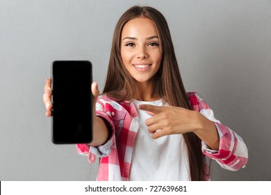 Young pretty smiling woman pointing with finger on phone screen, over gray wall