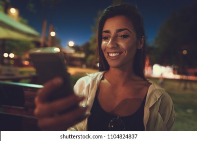 Young pretty, smiling woman surfing net with smartphone at evening time on the river bank. Selective focus.