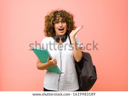young pretty student woman looking happy and excited, shocked with an unexpected surprise with both hands open next to face against pink wall