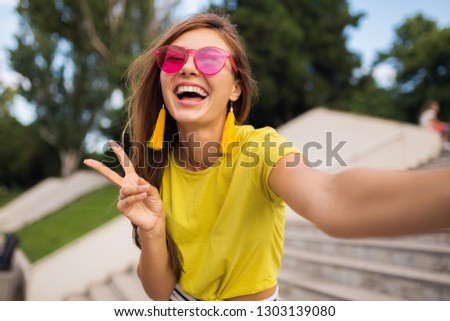 young pretty stylish smiling woman making selfie photo on camera in city park, positive, emotional, wearing yellow top, pink sunglasses, summer style fashion trend