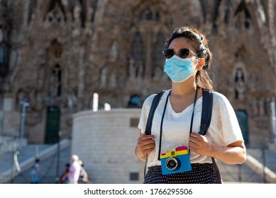 Young And Pretty Tourist Woman With Face Mask Posing In Front Of Sagrada Familia In Barcelona, Symbol Of Tourism To Spain During The Covid Disease And Corona Virus Pandemic