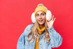 Young Pretty Uruguayan Woman Wearing Winter Muffs Isolated On Red Background Background Listening To Something By Putting Hand On The Ear