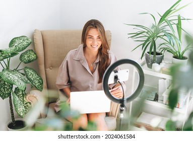 Young pretty woman blogger in records video on mobile phone using ring lamp sitting on chair with laptop in room with green plants at the home - Powered by Shutterstock