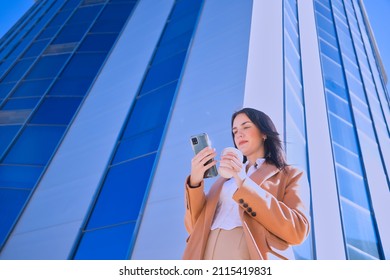 Young, Pretty Woman In Brown Coat And Glass Of Coffee In Hand, Checking Social Networks On Her Cell Phone On Her Way Out Of The Office. Counterpoint Concept, Business, Mobile, Working, Empowerment.