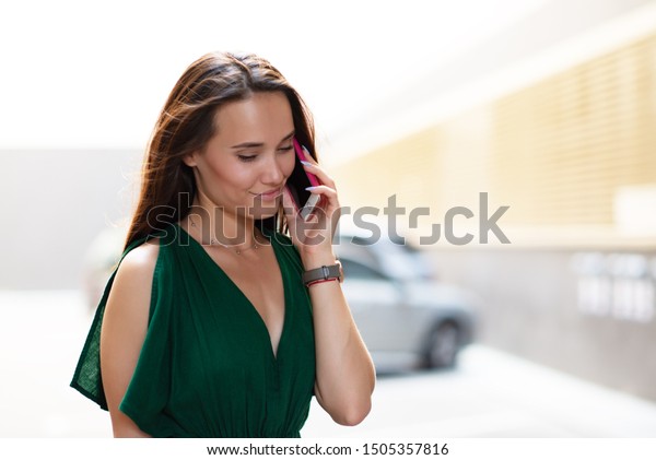 Young pretty woman calling\
phone, summer city outdoor. Beautiful girl dressed in\
emerald-colored jumpsuit talking on a smartphone in house parking.\
Urban lifestyle