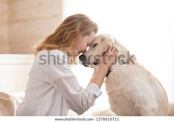 Young pretty woman in casual
clothes hugging her beloved big white dog sitting on the sofa in
the living room of her cozy country house. Animal communication
concept