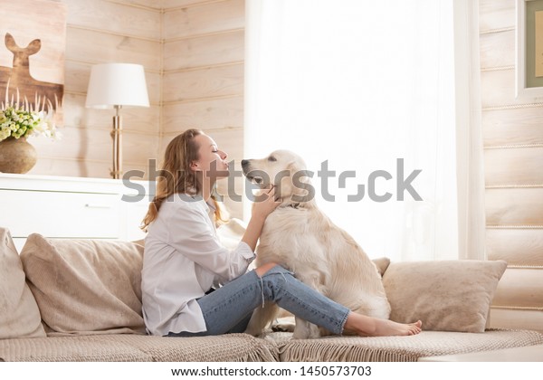 Young pretty woman in casual
clothes hugging her beloved big white dog sitting on the sofa in
the living room of her cozy country house. Animal communication
concept