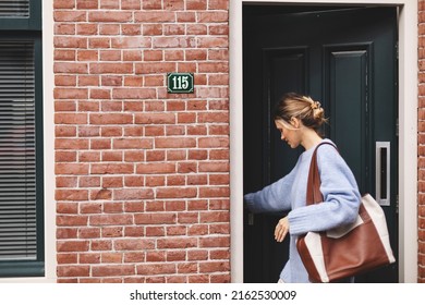 Young pretty woman in casual outfit going out of house. Girl wear blue sweater, brown bag and low bun on her hair and closed door of house. Woman go to market, work or meeting with friends.