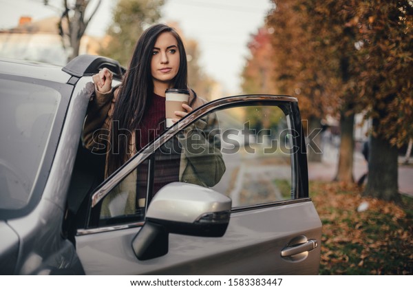 Young pretty woman with cup of\
coffee and texting blog on tablet in autumn park near\
vehicle