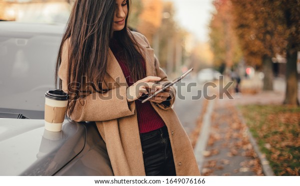Young pretty woman with cup of\
coffee and texting blog on tablet in autumn park near\
vehicle
