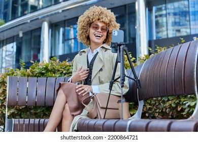 Young Pretty Woman With Curly Blonde Hair Records Video On Smartphone Settled On Tripod Chats Online Uses Phone Webcam Laughs Joyfully Dressed In Stylish Clothes Sits On Wooden Bench Outside